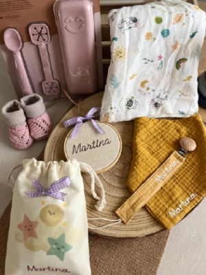 Conjunto de productos personalizados con el nombre "Martina", que incluye una bolsita de tela con dibujos de estrellas, una decoración circular con lazo lila, un babero mostaza, una pinza de chupete, una muselina blanca con dibujos infantiles y un par de zapatitos de punto rosados.