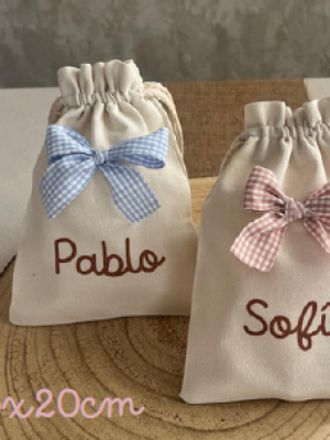 Dos bolsitas de tela color beige con cierre de cordón, decoradas con lazos de cuadros celeste y rosa. Están personalizadas con los nombres "Pablo" y "Sofía" en letras marrones. En la esquina se indica el tamaño “15x20cm”.