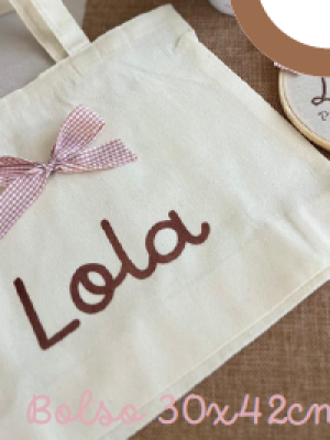 Bolsa de tela color beige con asas, personalizada con el nombre "Lola" en letras marrones y decorada con un lazo de cuadros rosado. En la esquina se lee “Bolso 30x42cm”.